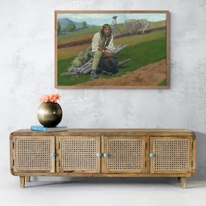 May include: A framed painting shows a man resting on logs in a field, with a farmer plowing in the background. Below, a wooden cabinet with woven doors and turquoise knobs holds a gold vase with pink flowers and a blue book.