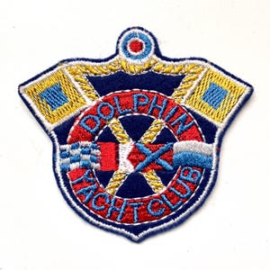 May include: Embroidered patch with a blue background and white outline. The center features a red and white nautical wheel with a yellow rope design. The text "DOLPHIN YACHT CLUB" is embroidered in white around the wheel. The patch has two yellow flags with blue squares on either side of the wheel.