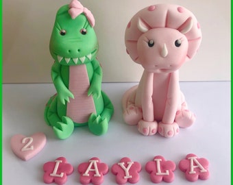 May include: A green dinosaur and a pink triceratops cake topper with a pink bow on the dinosaur's head. The cake topper is decorated with pink flowers and the name "LAYLA" in pink letters. There is also a pink heart with the number "2" on it.