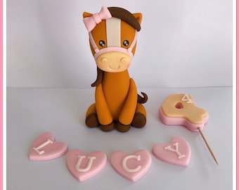 May include: A brown horse cake topper with a pink bow and bridle, next to a number four cake topper and pink heart-shaped letters spelling "LUCY". The horse is made of fondant and is a cute, whimsical design.