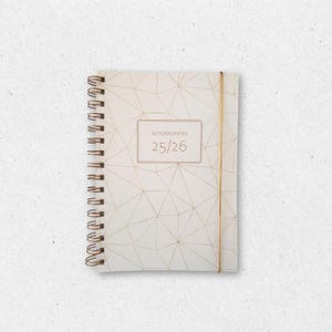 May include: A white spiral-bound notebook with a gold geometric pattern. The cover has a rectangular label with the text "Schülerplaner 25/26". A gold elastic band is wrapped around the notebook.