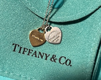 May include: A silver chain necklace with two heart-shaped pendants. The larger pendant is silver and says "PLEASE RETURN TO TIFFANY & CO. NEW YORK 925". The smaller pendant is gold and says "T&CO 750". The necklace is in a turquoise velvet pouch with the Tiffany & Co. logo.