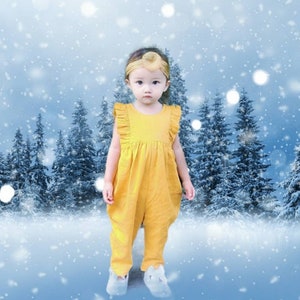 May include: A young child wearing a yellow ruffled jumpsuit stands in a snowy winter scene. The child is looking at the camera with a serious expression.