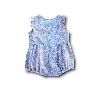May include: A white baby onesie with a floral print and a blue button. The onesie has ruffles on the shoulders.