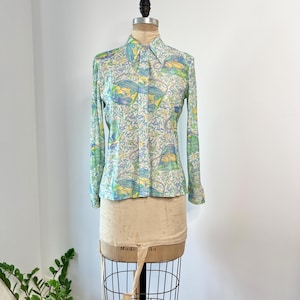 Könnte beinhalten: Ein hellblaues, langärmliges Hemd mit Knöpfen und einem bunten Blumenmuster. Der Druck zeigt grüne, gelbe und blaue Blumen und Blätter.