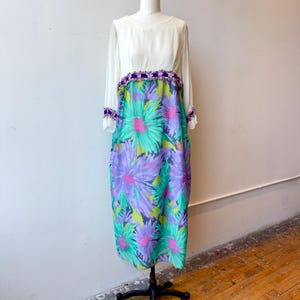 May include: A long-sleeved dress featuring a solid white bodice and a skirt with a vibrant floral pattern in shades of purple, teal, and lime green. A decorative trim of small purple flowers accents the waistline and the cuffs of the sleeves.
