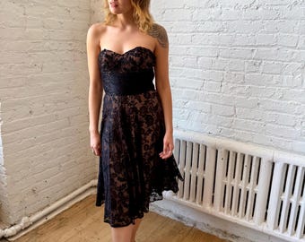 May include: A strapless black lace dress with a sweetheart neckline and a black ribbon belt. The dress has a knee-length hemline and is worn with black flats. The dress is set against a white brick wall and wooden floor.