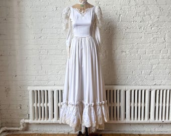 Op de afbeelding: Een witte vintage trouwjurk met lange mouwen en een volle rok. De jurk heeft kanten details op de mouwen, halslijn en zoom. Het wordt getoond op een zwarte paspop tegen een witte bakstenen muur.