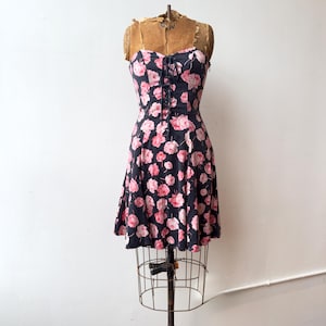 Könnte beinhalten: Ein schwarzes Vintage-Kleid mit rosa Blumenmuster, präsentiert auf einer Vintage-Schneiderpuppe. Das Kleid hat ein eng anliegendes Oberteil mit Schnürdetail und einen ausgestellten Rock. Das Kleid ist ärmellos und hat einen herzförmigen Ausschnitt.