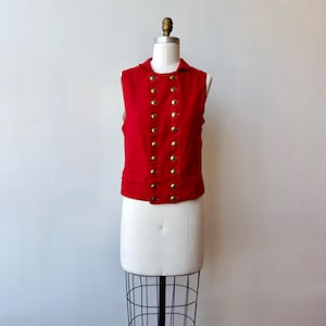 May include: A sleeveless red vest featuring a double-breasted front closure with two vertical rows of round, metallic gold-toned buttons. The garment has a structured collar, a straight hemline, and two small horizontal pockets at the lower front.