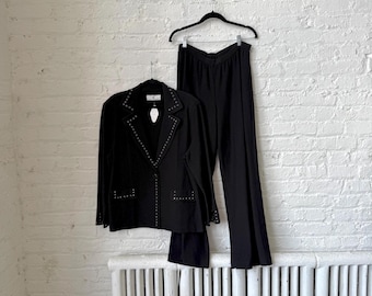 May include: A black two-piece suit hanging against a white brick wall. The suit includes a blazer with studded accents and matching wide-leg pants. The blazer is buttoned and has a notched lapel. The pants are high-waisted and have a flowing silhouette.