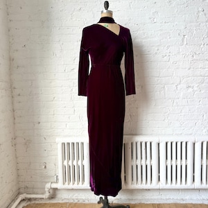 Könnte beinhalten: Ein langes, bordeauxfarbenes Samtkleid mit langen Ärmeln und einem einzigartigen Ausschnitt. Das Kleid wird auf einer Schaufensterpuppe vor einer weißen Backsteinmauer präsentiert. Das Kleid hat eine taillierte Taille und einen bodenlangen Rock.