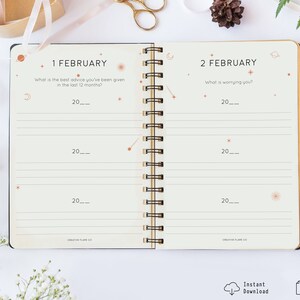 May include: A printable PDF journal with a cream-colored background and black spiral binding. The journal has a celestial theme with stars, planets, and moons. The text on the pages reads "1 February" and "What is the best advice you've been given in the last 12 months?" and "2 February" and "What is worrying you?"