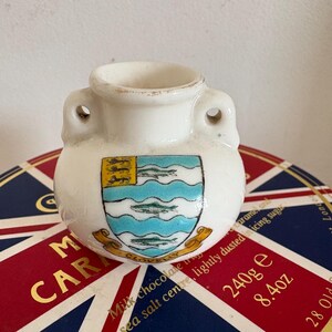 Puede incluir: Un pequeño tarro de cerámica blanca con dos asas. El tarro tiene un escudo azul y verde con fondo amarillo y las palabras "CLOVELLY" impresas en él. El tarro está sentado encima de una caja roja, blanca y azul con las palabras "Milk chocolate caramel" y "The Royal Arcade" impresas en ella.