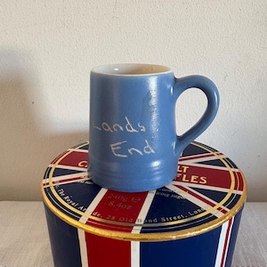 Może przedstawiać: Jasnoniebieski kubek ceramiczny z napisem "Lands End". Kubek stoi na niebiesko-czerwono-białym pudełku z złotymi akcentami.