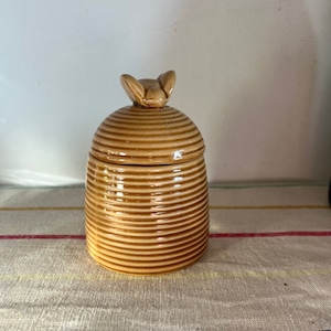 May include: A ceramic honey pot with a bee-shaped lid. The pot is brown and has a striped pattern.