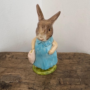 Puede incluir: Figura de cerámica de un conejo con un vestido azul y una bolsa rosa. El conejo tiene pelaje marrón y está sobre una base verde. La figura está sobre un fondo neutro.