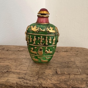 Puede incluir: Una botella decorativa verde con detalles dorados y un tapón rojo. La botella presenta diseños dorados en relieve de animales y jeroglíficos. La botella se exhibe sobre una superficie de madera rústica.