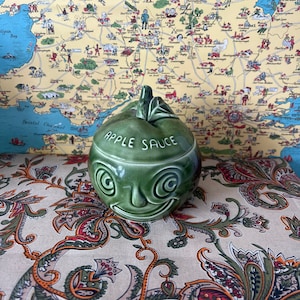May include: A green ceramic apple-shaped container with a lid. The container has a smiling face with swirling eyes and the words "APPLE SAUCE" written on the front.