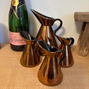 May include: Four copper-colored pitchers with black handles and spouts are arranged in front of a green champagne bottle. The pitchers vary in size, with a shiny, reflective surface. The champagne bottle has a pink label.