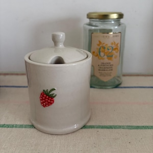 May include: A white ceramic sugar bowl with a lid and a small red strawberry design. The bowl is sitting on a green and white striped surface.