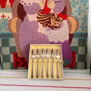 May include: A boxed set of six silver forks. The forks have a decorative handle design. The background features a tapestry depicting a woman knitting with a cat. The tapestry is in shades of purple, green, and orange.