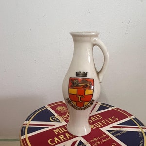 Puede incluir: Una jarra de cerámica blanca con asa, con un escudo de armas colorido con un león y una corona. La jarra está sobre una caja roja, blanca y azul con el texto "Milk Chocolate Truffles with Caramel and Sea Salt Centres, Lightly Dusted Icing Sugar, 240g e, 8.4oz, One, The Royal Arcade, 28 Old Bond Street, London".