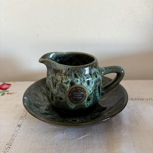 May include: A green and white ceramic creamer and saucer set with a gold sticker that reads "Foster Studio Pottery, Made in the West Country".