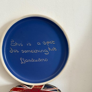 Op de afbeelding: Een blauwe keramische plaat met een witte rand, met de tekst "This is a spot for something hot Llandudno". Daaronder een cilindervormige blik met een Union Jack-ontwerp en gouden rand.