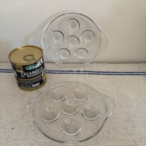 May include: Two clear glass escargot plates with six circular indentations each, and two handles. A can of escargots is next to one plate. The can has a gold lid and a label with text in French.