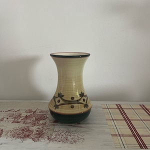 Puede incluir: Un jarrón decorativo con cuerpo color crema, base verde oscuro y borde marrón. El jarrón presenta un patrón de hojas verdes oscuras y puntos marrones. El jarrón mide aproximadamente 20 cm de altura.