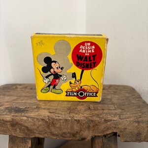 May include: Vintage yellow box featuring Mickey Mouse and Pluto from Walt Disney. The box has the text "UN DESSIN ANIME de WALT DISNEY" in a red circle. The box is sitting on a rustic wooden stool.