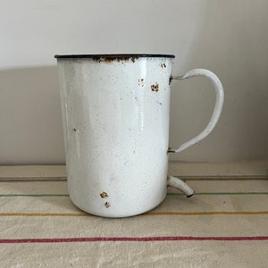Puede incluir: Jarra de esmalte blanco con borde negro y asa. La jarra tiene un pico y muestra signos de óxido.