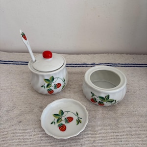 Könnte beinhalten: Ein weißes Keramik-Zuckerdosen-Set mit Deckel, Löffel und kleiner Schale. Das Set ist mit roten Erdbeeren und grünen Blättern verziert. Der Deckel hat einen roten Knopf. Der Löffel hat ein Erdbeermuster.