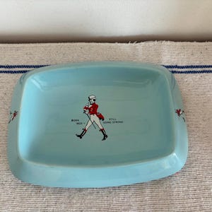 May include: A light blue rectangular dish with rounded corners. The dish features a graphic of a man in a red coat and the text "BORN 1820 - STILL GOING STRONG". Decorative accents are on the sides of the dish.