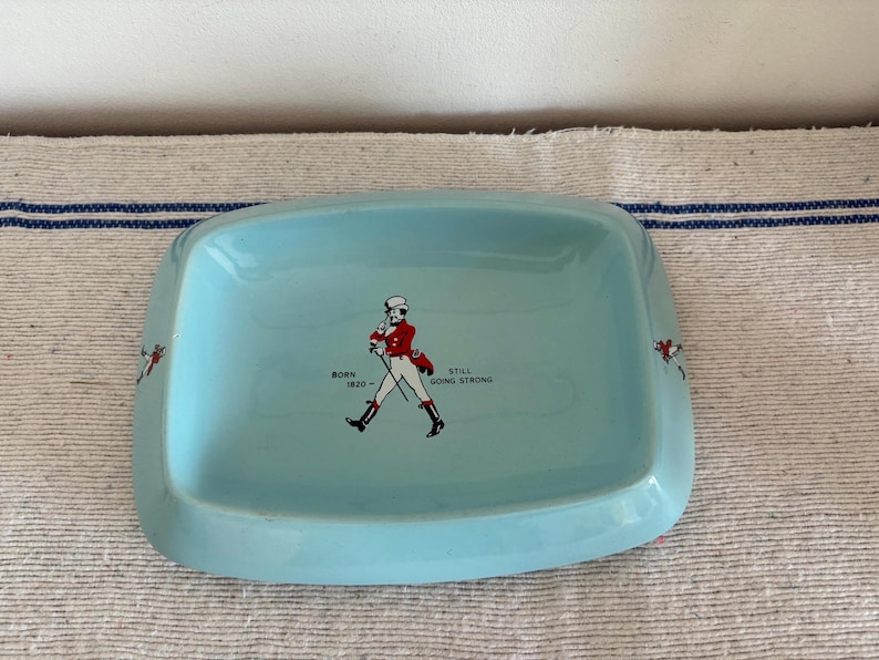 May include: A light blue rectangular dish with rounded corners. The dish features a graphic of a man in a red coat and the text "BORN 1820 - STILL GOING STRONG". Decorative accents are on the sides of the dish.