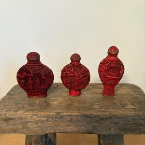 Puede incluir: Tres botellas de rapé rojas antiguas talladas con intrincados diseños en relieve. Cada botella presenta una escena detallada y una tapa de color latón. Las botellas se exhiben sobre un taburete de madera rústica, mostrando su artesanía ornamentada.