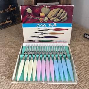 Op de afbeelding: Een set van twaalf roestvrijstalen cocktailvorkjes met kleurrijke kunststof handvatten in wit, geel, roze en blauw, gerangschikt in een kartonnen doos. Het deksel bevat de tekst Little Fork en Stainless Steel met een afbeelding van fruit.
