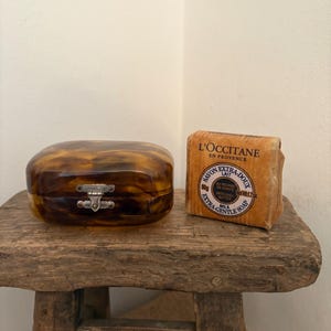 Pode incluir: Uma caixa com padrão de tartaruga e fecho prateado está ao lado de uma barra de sabonete de leite Extra-Gentle L'Occitane. O sabonete está numa embalagem de papel castanho com texto em francês. Ambos os itens estão numa pequena banqueta de madeira rústica.