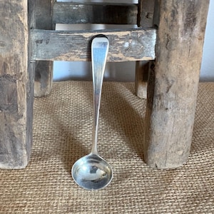 May include: A small, silver ladle with a rounded bowl and long handle rests on a textured, tan surface. The ladle is positioned in front of a rustic, wooden stool with visible grain and aged appearance. The background is neutral.