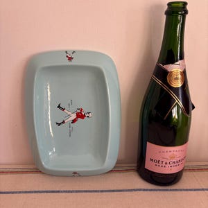 May include: A light blue ceramic ashtray with a red and white illustration of a person riding a horse. The ashtray is next to a bottle of Mo&euml;t & Chandon Ros&eacute; Imp&eacute;rial champagne.