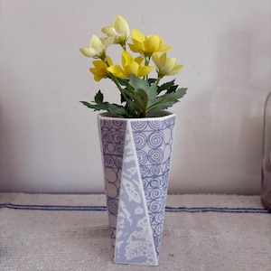 May include: A patterned vase in blue and white, containing yellow flowers and green foliage. The vase has a distinctive shape and is set on a patterned surface. The vase is approximately 20 cm tall.
