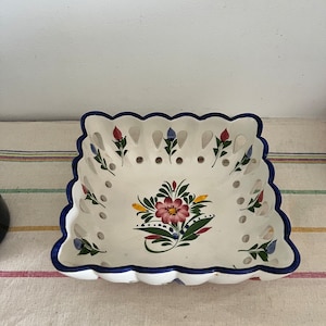 May include: White ceramic square bowl with a blue and white scalloped edge. The bowl has a floral design in pink, yellow, green, and blue. The bowl has a lattice design with holes throughout.