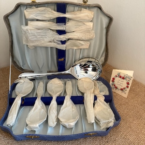May include: Vintage silver-plated spoon set in a blue and gray case. The set includes a serving spoon and several smaller spoons, all wrapped in tissue paper. The case has a blue satin lining and a decorative clasp.