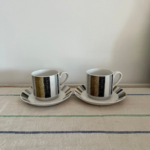 May include: Two white ceramic teacups with black, gold, and grey striped designs. The cups are sitting on matching saucers on a striped tablecloth.