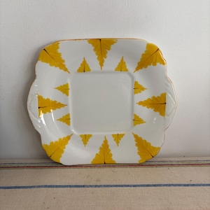 May include: A white square ceramic plate with a decorative border of yellow fern leaves. The plate has a scalloped edge and two small handles.