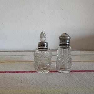 Może przedstawiać: Dwa zabytkowe szklane flakony perfum ze srebrnymi metalowymi nakrętkami. Butelka po lewej stronie ma fasetowany korek. Butelka po prawej stronie ma perforowaną nakrętkę. Obie butelki są przezroczyste i stoją na paskowanej tkaninie.