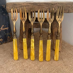 Peut inclure: Six fourchettes antiques avec des dents et des manches en laiton. Les manches sont de couleur jaune marbré. Les fourchettes sont disposées en rangée sur une surface en bois rustique, suggérant un thème vintage ou de collection.