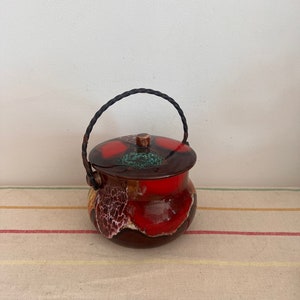 May include: A small, red and brown ceramic jar with a lid and a black braided handle. The jar has a mottled glaze with white and green accents.