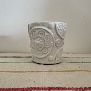 Könnte beinhalten: Weißer Keramiktopf mit einem erhabenen Blumendesign. Der Topf hat eine abgerundete Form und eine leicht strukturierte Oberfläche.